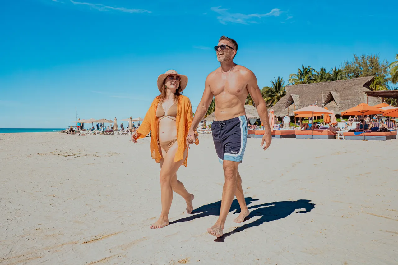 Pareja adulta joven caminando tomados de la mano en una playa de arena blanca con aguas turquesas al fondo. Al lado derecho se ve un club de playa con palapas con sombrillas naranjas, camastros y restaurante. Ambiente relajado y vacacional de día soleado.