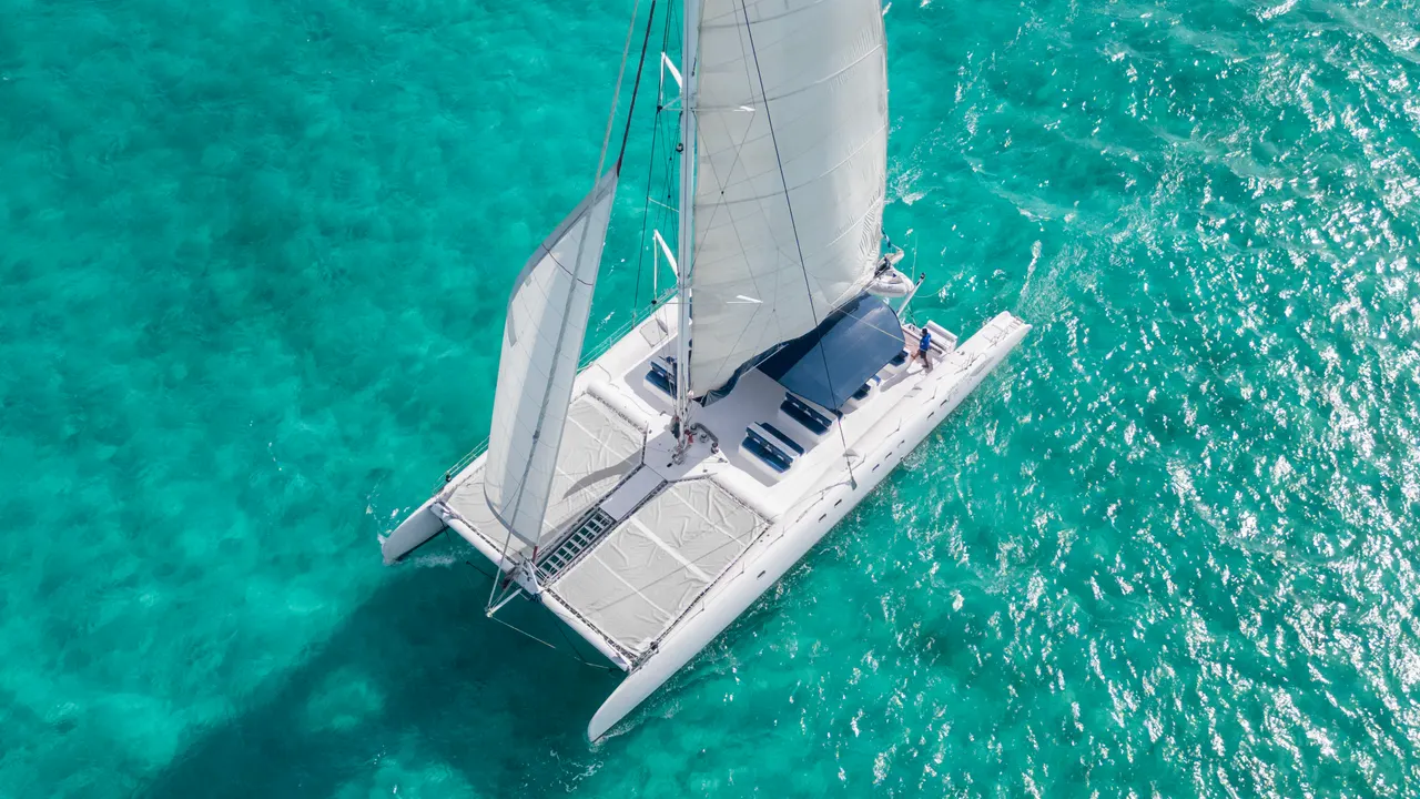 Vista aérea de un catamarán blanco de lujo navegando en aguas cristalinas turquesas del Caribe. La toma cenital muestra el barco con velas desplegadas, cubierta despejada y amarras visibles, rodeado de mar de color azul intenso. Captura la elegancia y serenidad de un día de navegación perfecto.