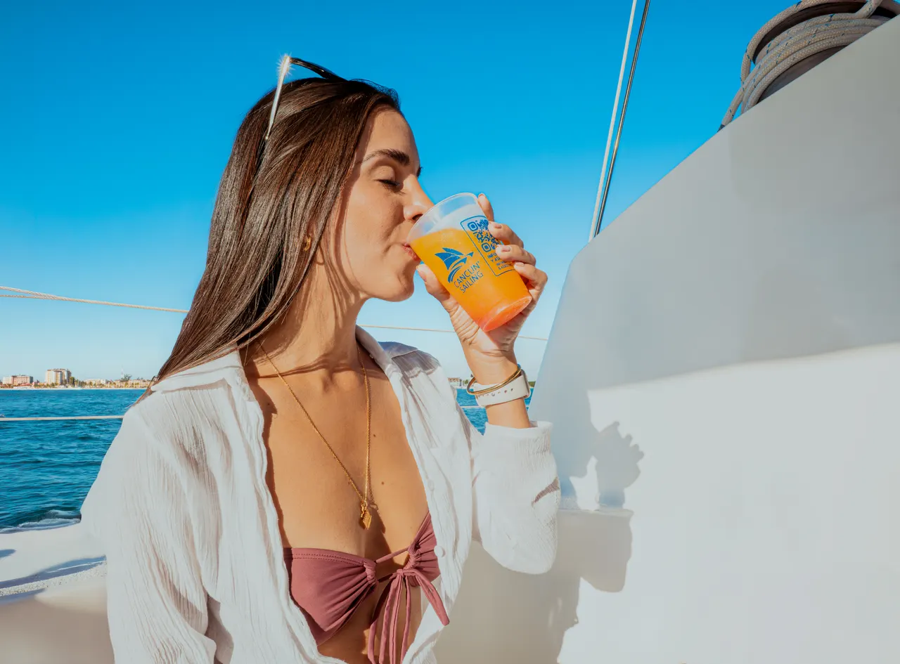 Mujer joven en traje de baño con camisa blanca semitransparente bebiendo un refresco naranja en la cubierta de un catamarán. El mar turquesa y la costa de Cancún se ven al fondo bajo un cielo despejado. Ambiente relajado y casual, ideal para promocionar la experiencia de ocio en el mar.