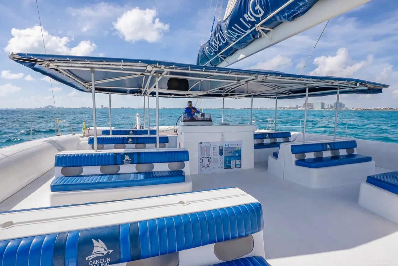 Vista de la cubierta principal de un catamarán Cancun Sailing con amplios asientos acolchados en azul y blanco, toldo de vela azul desplegado, y un tripulante en la cabina de mando. Al fondo se observa el mar turquesa caribeño y la costa de Cancún bajo un cielo despejado con nubes blancas.