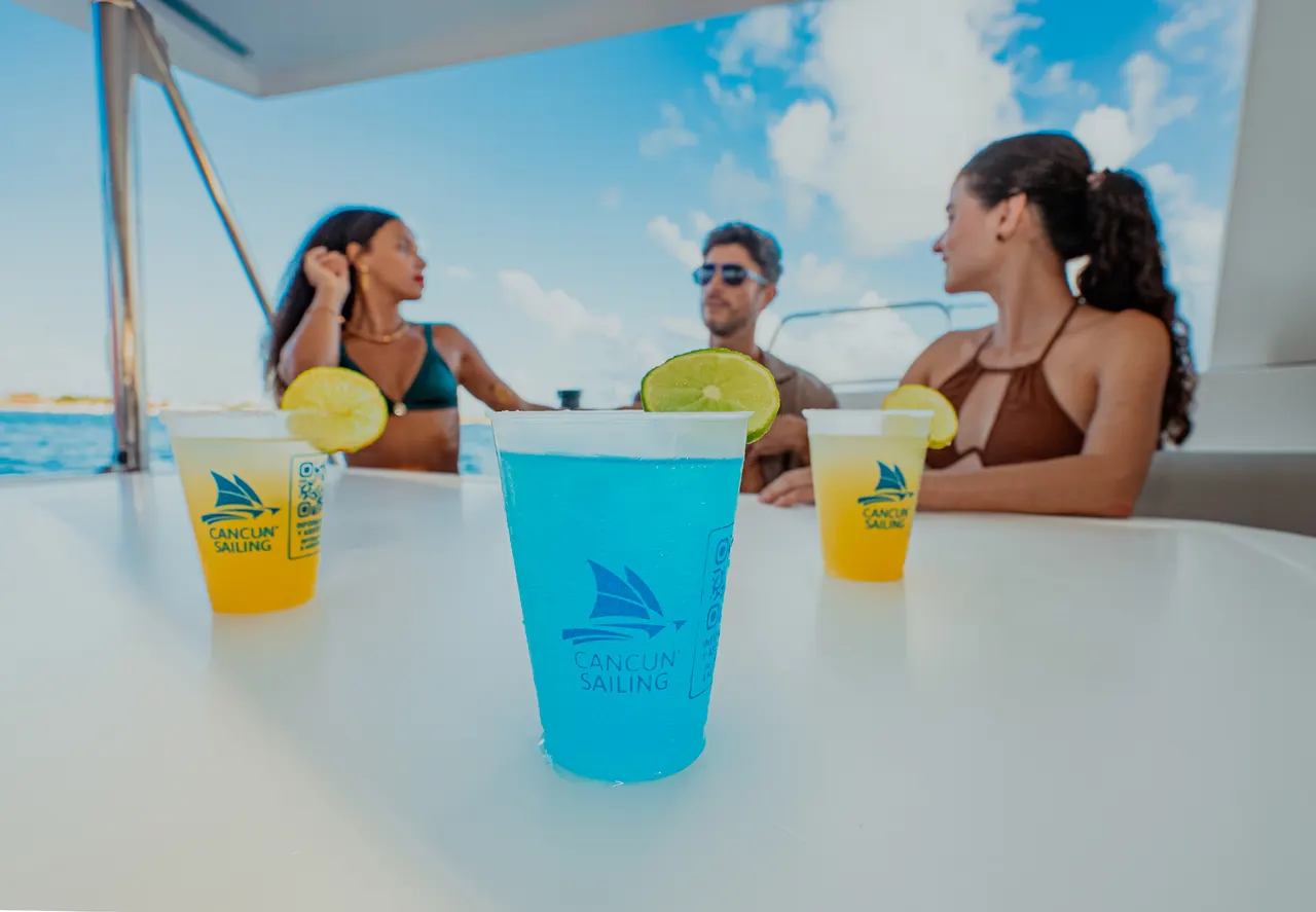 Grupo de amigos adultos brindando con cócteles tropicales en vasos con logo de Cancun Sailing, decorados con rodajas de limón. Ambiente alegre y desenfadado en la cubierta del catamarán, con el mar turquesa y cielo despejado de fondo. Perfecta para mostrar la experiencia social y refrescante de los tours.