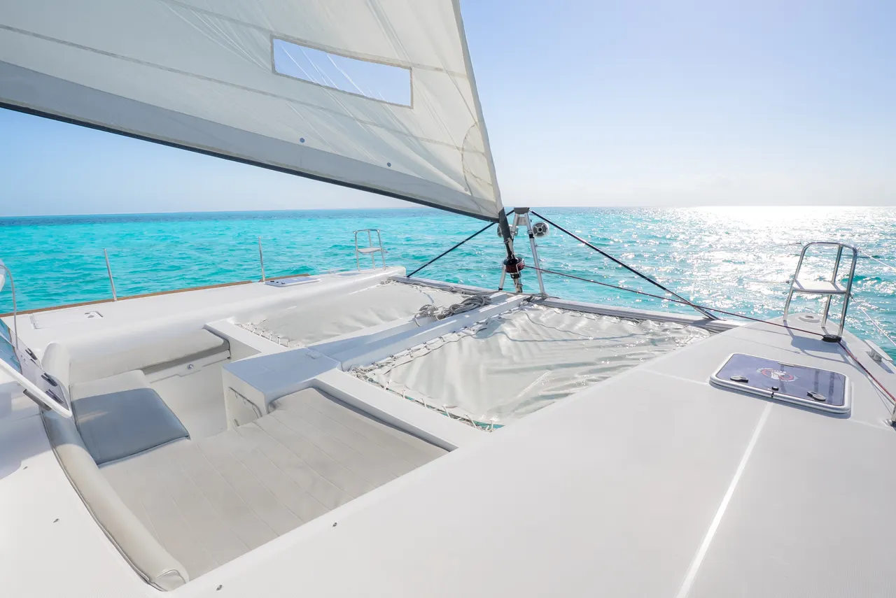 Vista de la cubierta de proa de un catamarán de lujo con vela blanca desplegada, mostrando la red de proa y áreas de descanso acolchadas. El agua turquesa del Caribe brilla al fondo bajo iluminación diurna, capturando la estética premium y navegabilidad de la embarcación.