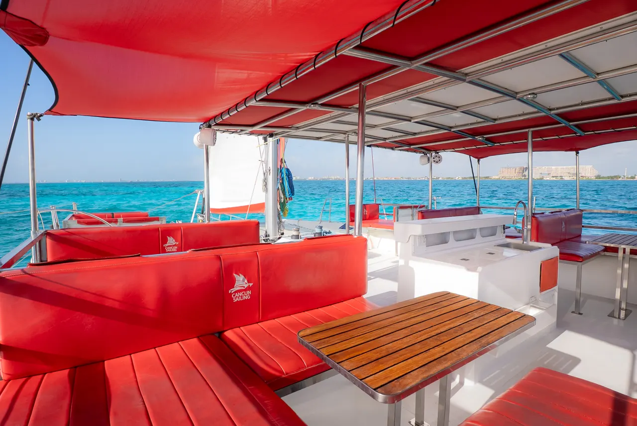 Vista de la cubierta principal de un catamarán Cancun Sailing equipado con amplios cojines rojos, toldo rojo retráctil y mesa de madera. El mar turquesa del Caribe se extiende al fondo con la costa de Cancún visible. Ambiente abierto, moderno y cómodo, ideal para relajarse durante el tour.