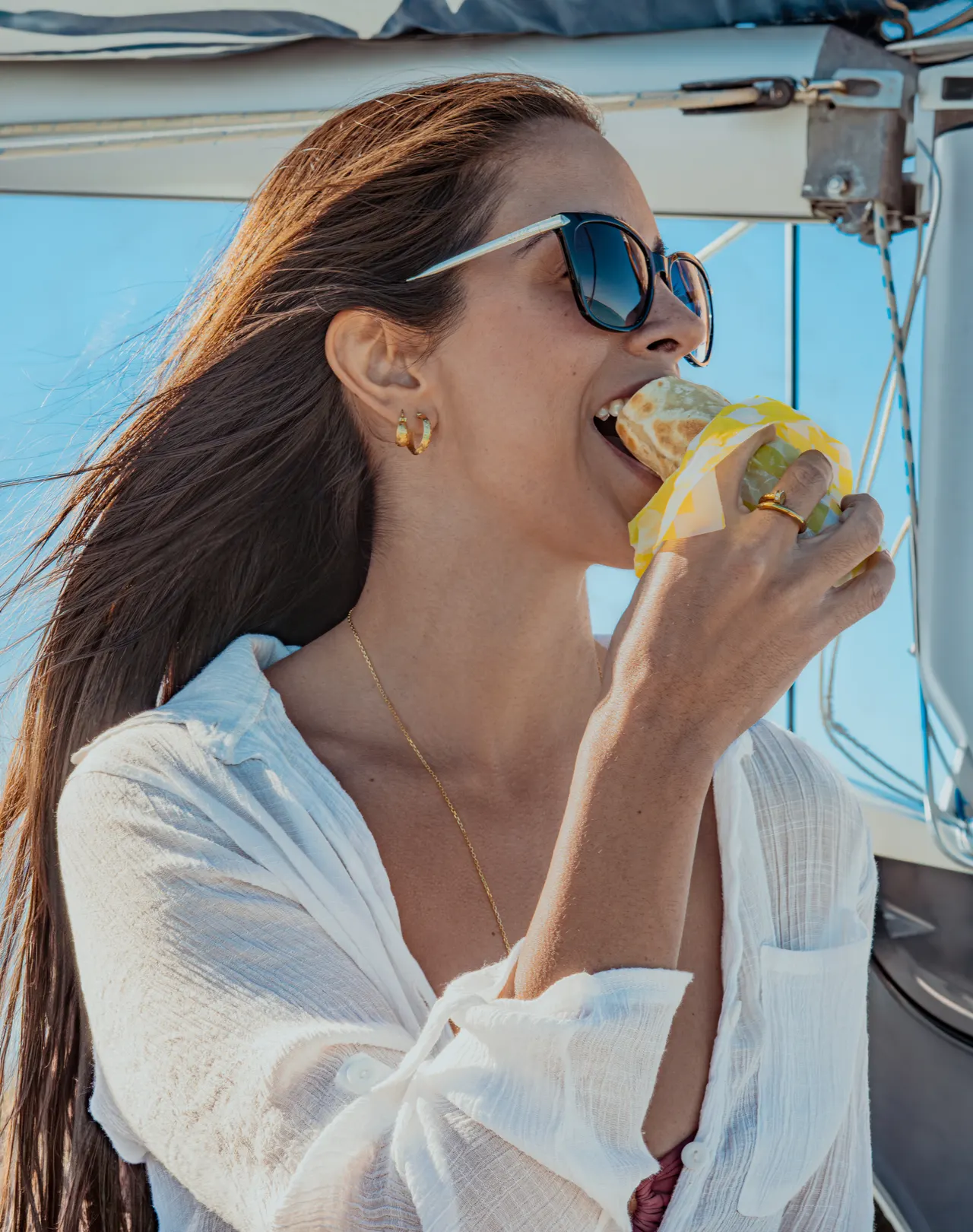 Mujer adulta sonriendo mientras disfruta de un sándwich envuelto en papel amarillo en la cubierta de un catamarán. Viste blusa blanca, lentes de sol, y está rodeada de cielo azul despejado. La fotografía captura un momento casual y relajado de gastronomía a bordo.