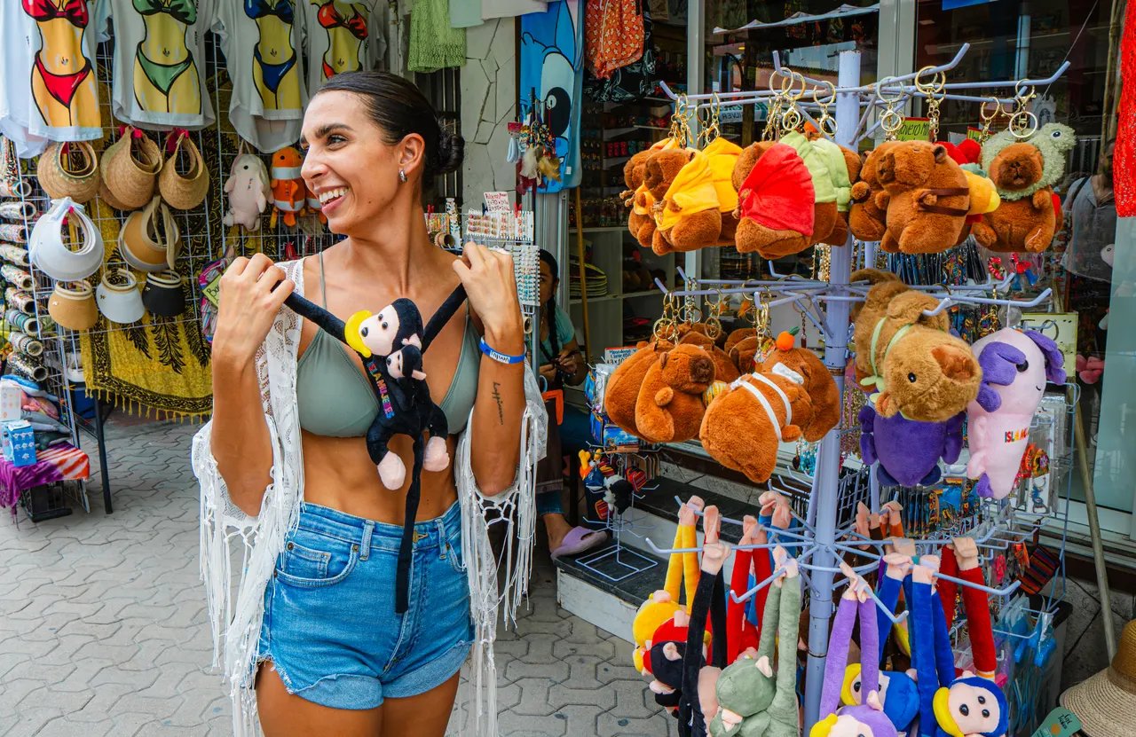 Mujer joven sonriente comprando souvenirs en una tienda colorida de Isla Mujeres, rodeada de sombreros, peluches y artesanías típicas. Ambiente vibrante y casual de shopping en la isla, reflejando la experiencia turística local.