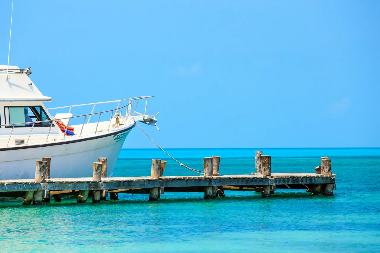 Lancha blanca amarrada a un muelle de madera desgastado en aguas cristalinas turquesas del Caribe. La embarcación muestra detalles de su proa y equipo de amarre bajo un cielo despejado, con el horizonte del mar infinito de fondo.