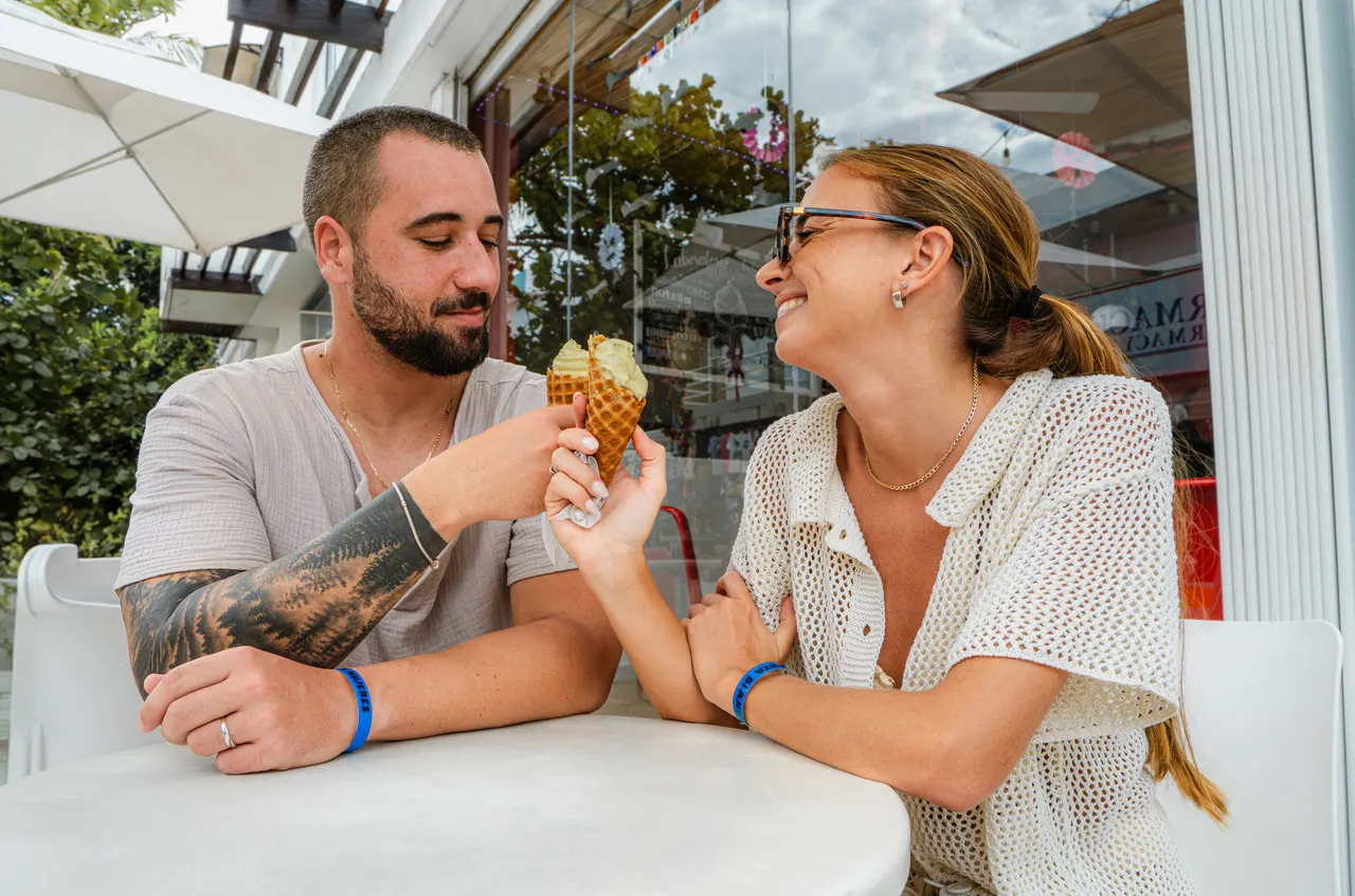 Pareja joven sonriendo mientras comparten un helado en una calle comercial de Isla Mujeres. Ambiente casual y romántico, con tiendas y vegetación tropical de fondo. Muestra el lado turístico y relajado de la isla, ideal para promocionar la experiencia completa del viaje.