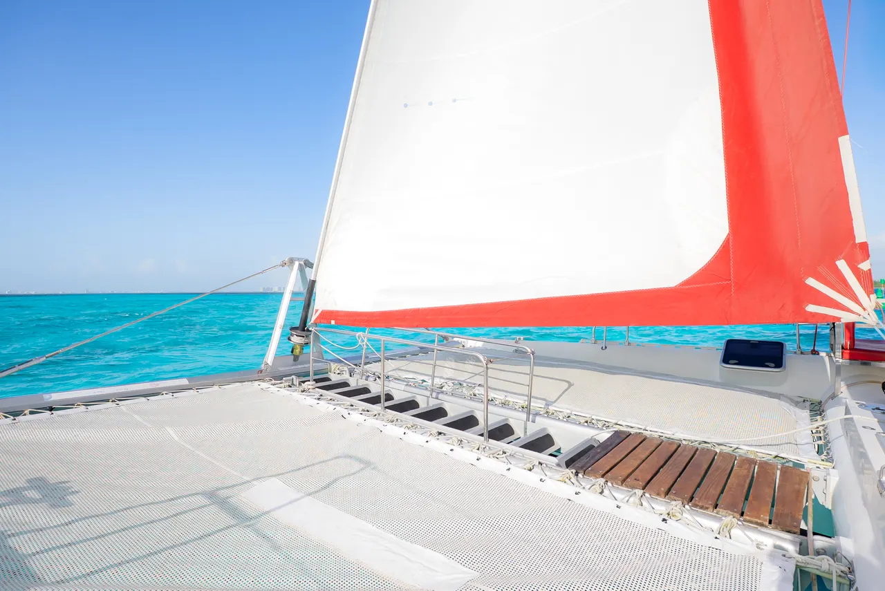 Vista de la cubierta frontal de un catamarán con velas blanca y roja desplegadas, navegando en aguas turquesas cristalinas del Caribe. La red de proa y detalles del barco son visibles bajo un cielo azul despejado, reflejando la elegancia y calidad de la embarcación.