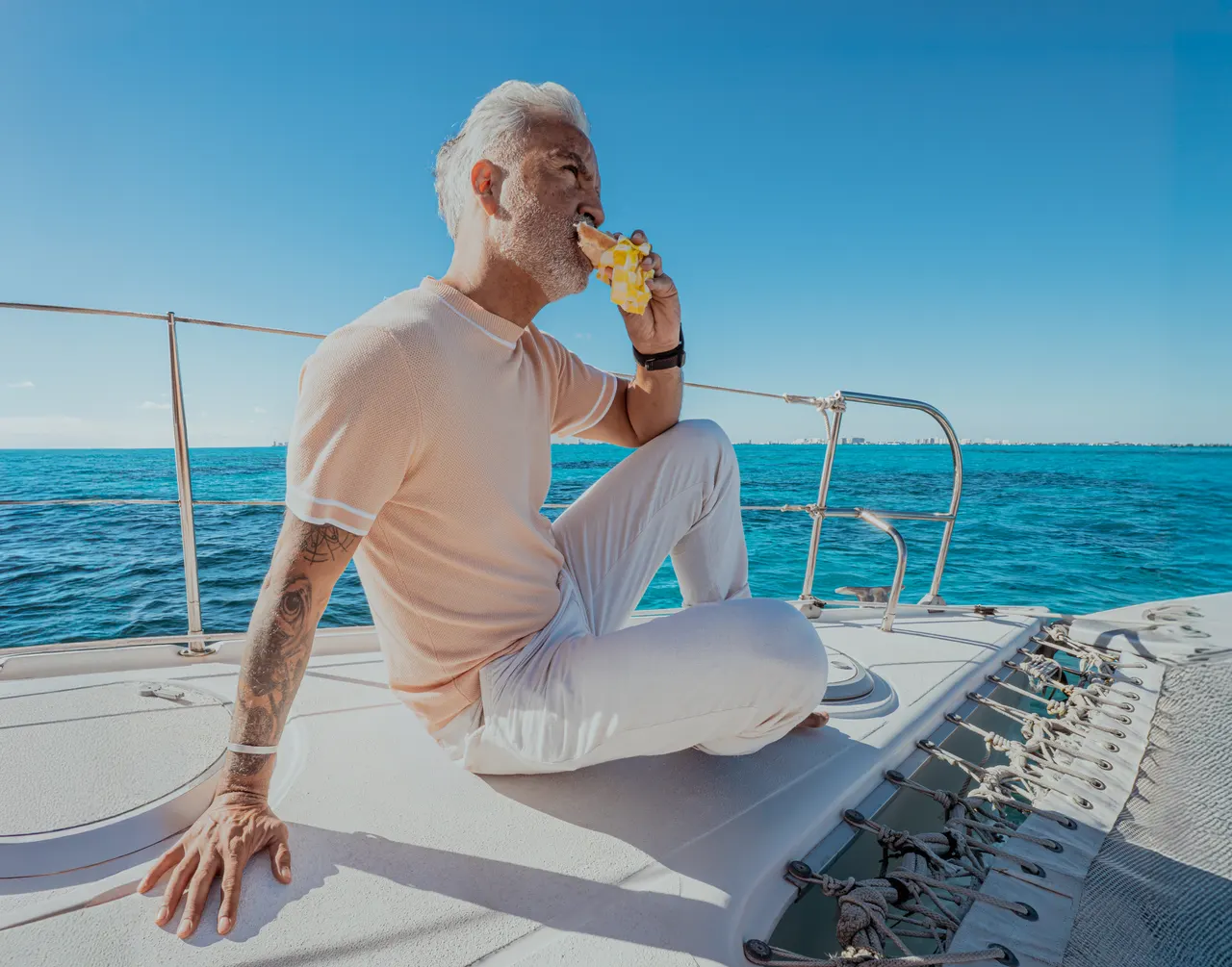 Hombre adulto mayor sentado en la cubierta de un catamarán comiendo piña fresca, con el mar turquesa del Caribe de fondo bajo un cielo despejado. Ambiente relajado y casual, mostrando el disfrute de la gastronomía a bordo durante el tour.