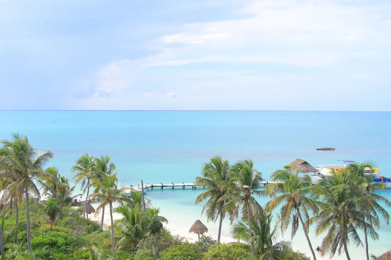 Vista panorámica aérea de Playa Norte en Isla Mujeres mostrando arena blanca, aguas turquesas del Caribe, palmeras cocos frondosas y un muelle de madera. El paisaje natural captura la belleza virgen de la isla con instalaciones mínimas, ideal para promocionar excursiones a Isla Mujeres.