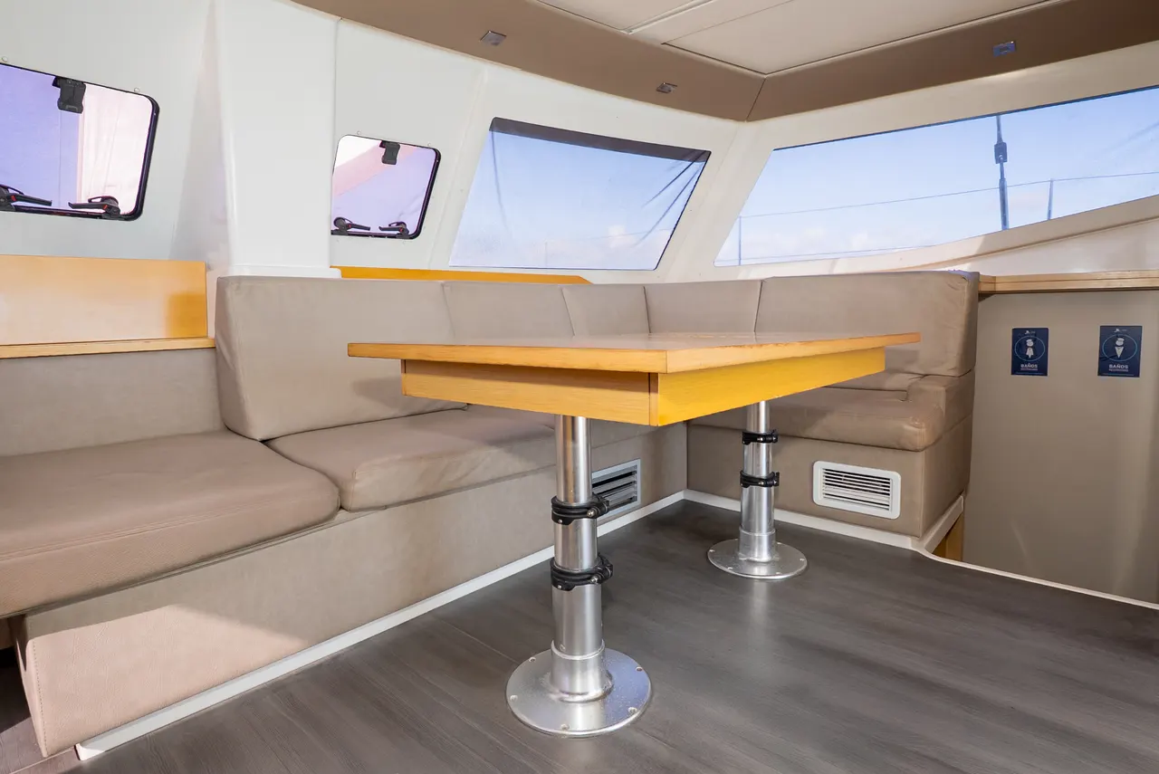 Interior espacioso de un catamarán de lujo mostrando una sala de estar con sofás beige, mesa central de madera amarilla con base plateada, grandes ventanales que ofrecen vistas al cielo azul y al mar. Diseño contemporáneo y premium con acabados en materiales de alta calidad.