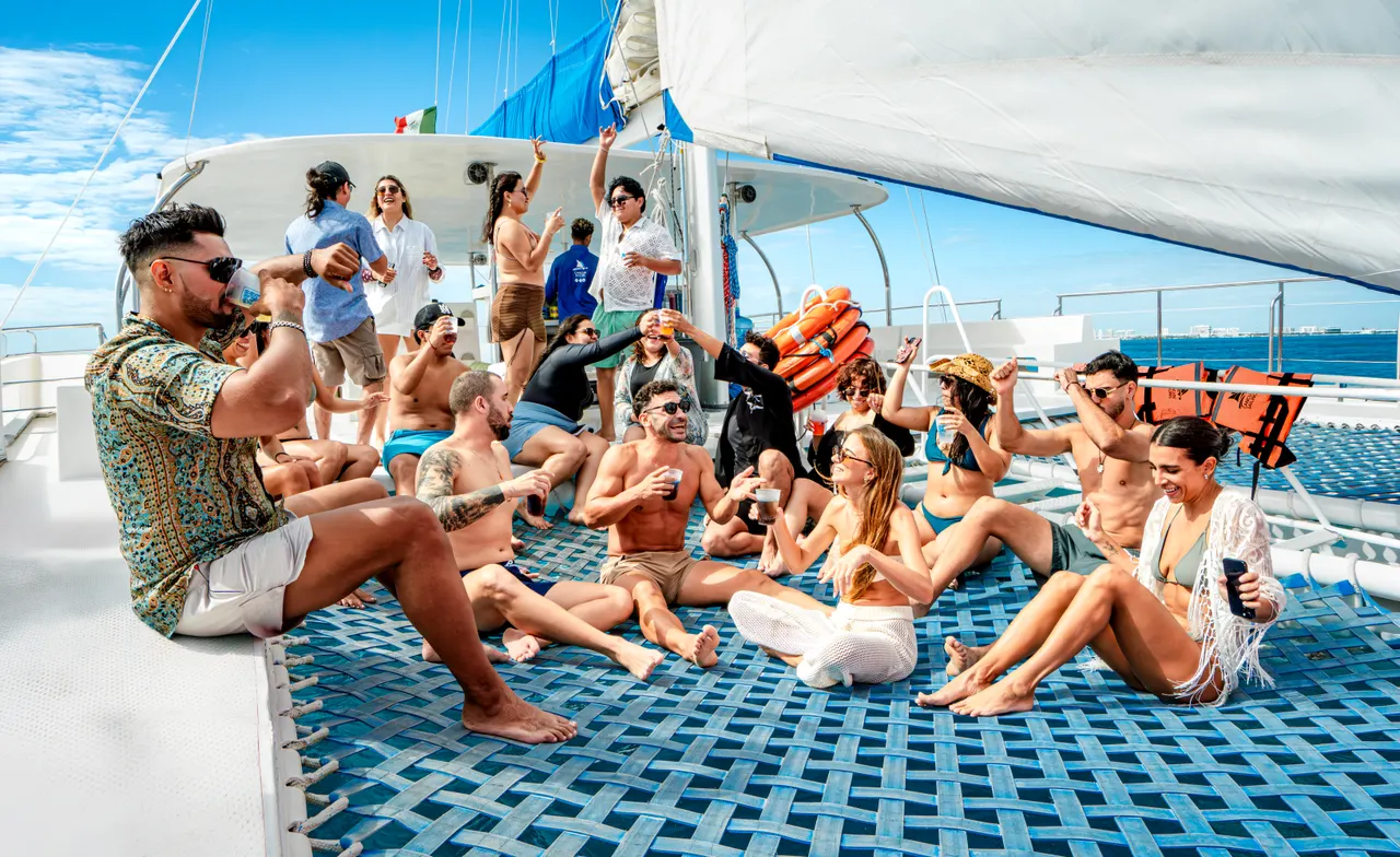 Grupo numeroso de amigos y turistas relajándose en la cubierta principal de un catamarán Privilege, con velas blancas desplegadas y el mar turquesa de fondo. Ambiente festivo y casual, con personas sentadas, tomando el sol y disfrutando de la navegación bajo un cielo despejado.