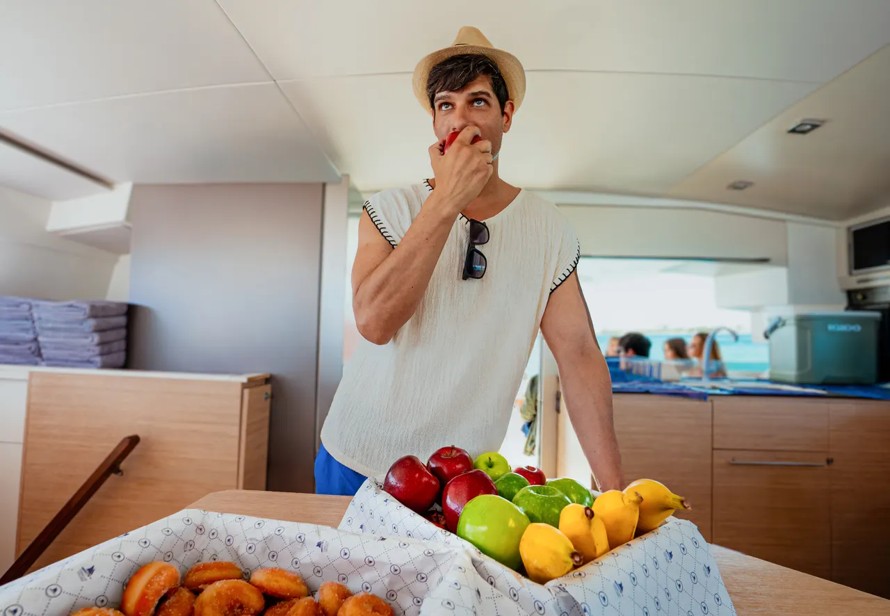 Hombre sonriente con sombrero de paja degustando fruta dentro de la cocina de un catamarán, rodeado de manzanas rojas, verdes y naranjas frescas. Al fondo se ven pasajeros en la cubierta disfrutando del mar turquesa. Ambiente casual y desenfadado que refleja la experiencia gastronómica abordo.