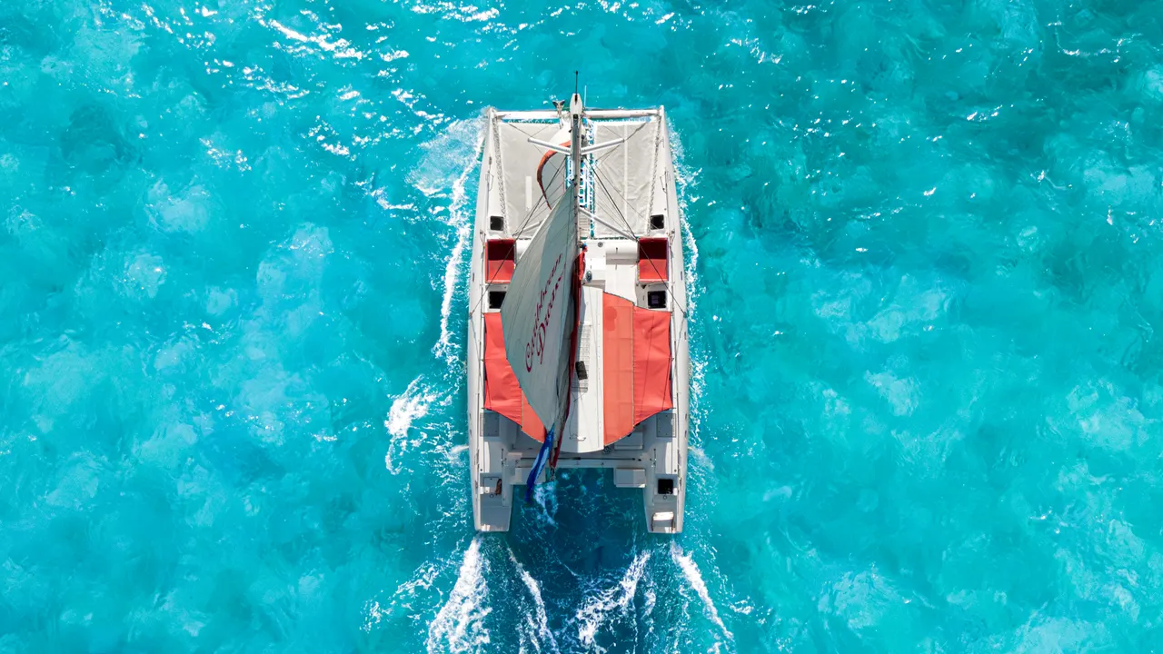 Vista aérea cenital de un catamarán blanco y rojo navegando en aguas cristalinas de color turquesa intenso. El barco crea una estela blanca característica mientras navega hacia el horizonte, mostrando toda la estructura del catamarán con sus dos cascos paralelos.
