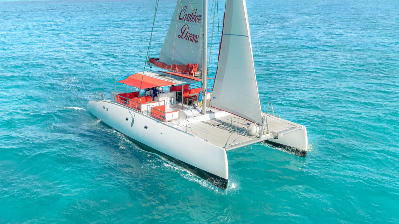 Catamarán blanco y rojo Caribbean Dreams en navegación sobre aguas turquesas del Caribe. Vista aérea que muestra la vela izadas, cubierta con toldo rojo y la estructura elegante del barco de dos cascos, capturando la belleza del mar cristalino.