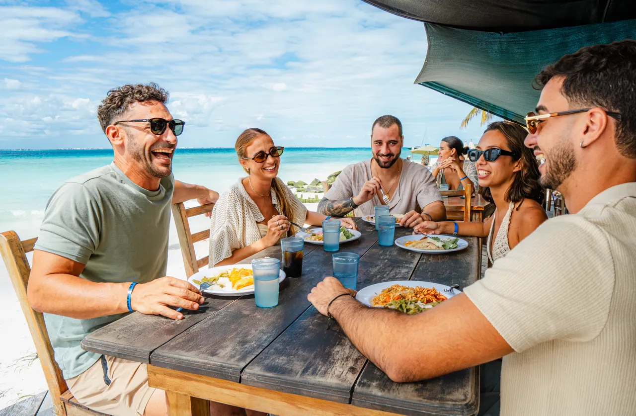 Grupo de seis adultos disfrutando de una comida al aire libre en un club de playa, sentados alrededor de una mesa de madera bajo una palapa. Al fondo se ve el agua turquesa del Caribe. Ambiente casual, relajado y festivo con todos sonriendo y compartiendo el momento.