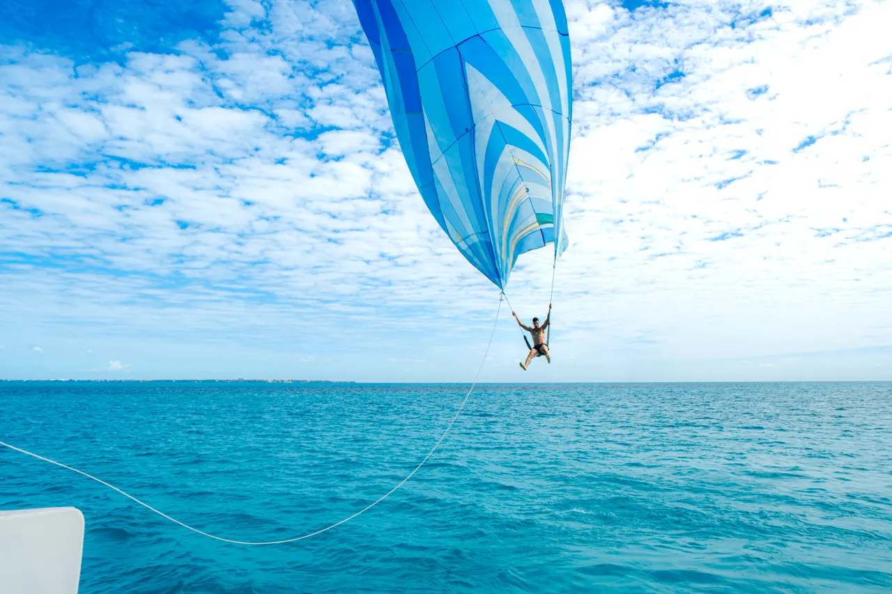 Persona suspendida en el aire realizando parasailing con una vela paracaídas azul y blanca desplegada, sobre aguas turquesas del Caribe. La foto captura la adrenalina y libertad de la actividad acuática, con cielo despejado y horizonte marino infinito.