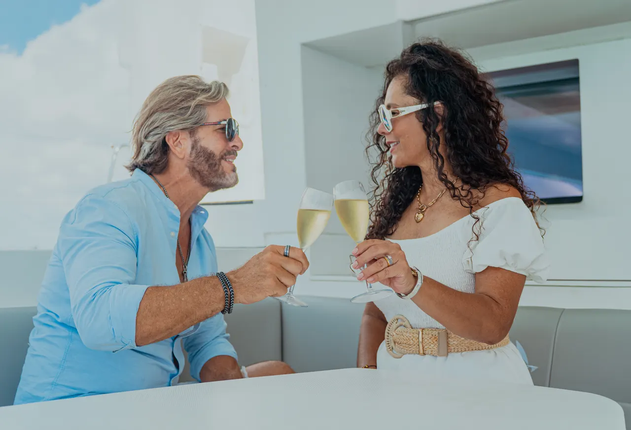 Pareja adulta en ambiente romántico brindando con copas de champagne blanco en la cubierta interior de un catamarán de lujo. Él viste camisa azul cielo y ella un vestido blanco con cinturón dorado, ambos con gafas de sol. El mar turquesa y el cielo azul se ven de fondo a través de las ventanas. Atmósfera exclusiva y celebratoria ideal para luna de miel o aniversario.