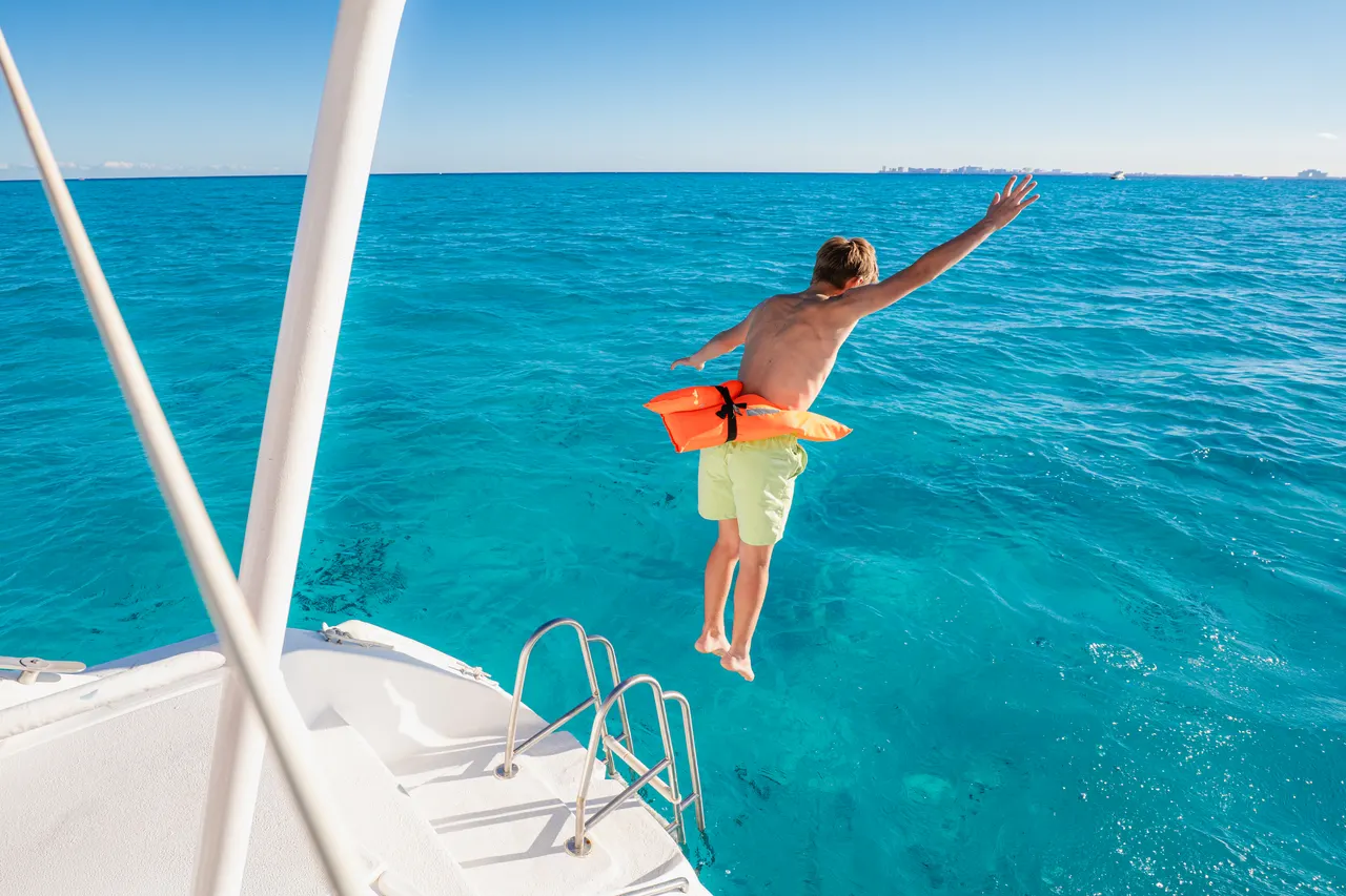 Niño pequeño saltando alegremente al agua turquesa del Caribe desde la cubierta de un catamarán, vistiendo chalecos salvavidas de color naranja. La escena captura la diversión y libertad de las actividades acuáticas en familia, con el agua cristalina y cielo azul de fondo.
