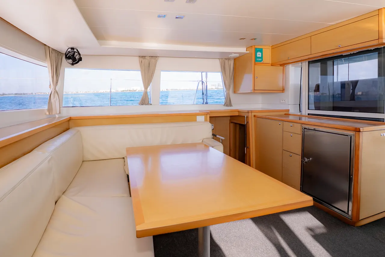 Interior espacioso de un catamarán de lujo mostrando la sala de estar con sofás de cuero crema, mesa de madera pulida, muebles de caoba y grandes ventanales que ofrecen vistas panorámicas del mar turquesa del Caribe. Ambiente refinado y confortable, ideal para viajes premium.