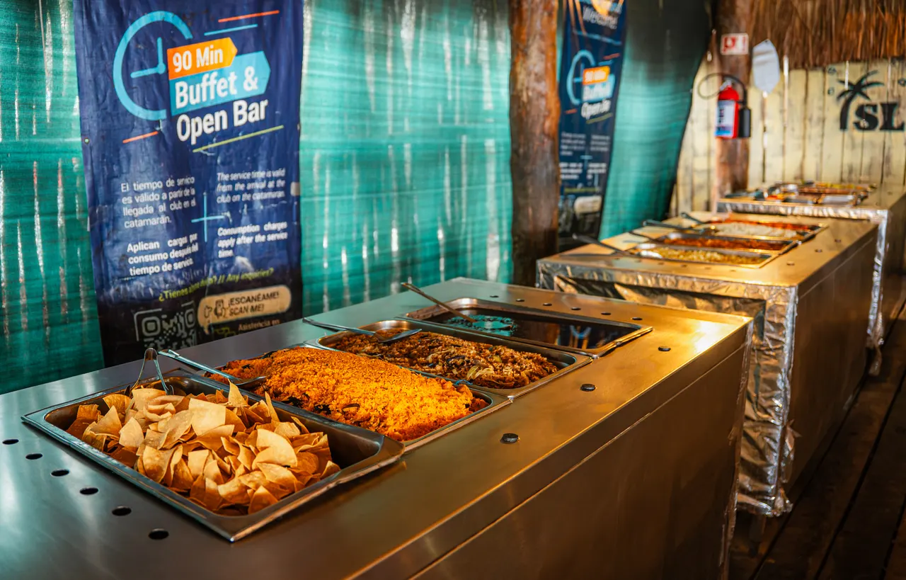 Buffet con platillos locales y botanas dispuestos en bandejas de servicio dentro de un club de playa. Se aprecia comida típica de la región con carnes sazonadas, chips y una barra de open bar. Ambiente de instalaciones gastronómicas de day-pass en Isla Mujeres.