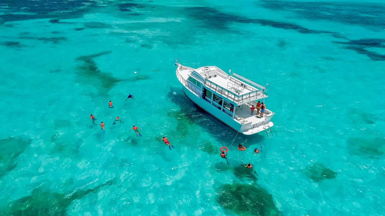 Vista aérea de un grupo de turistas en trajes de baño naranja esnorqueando en las aguas cristalinas y turquesas del Caribe, rodeados de vida marina visible bajo el agua. Un barco de tour está anclado cercano con tripulación a bordo, capturando la esencia de una excursión de snorkel en Isla Contoy.