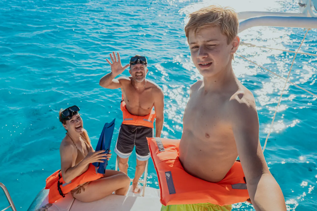 Familia con niños en el agua cristalina del Caribe, sonriendo y disfrutando de un chapuzón desde la cubierta de un catamarán. El agua turquesa intenso, los chalecos de seguridad naranjas y la atmósfera alegre reflejan un día perfecto en familia explorando las aguas de Cancún.