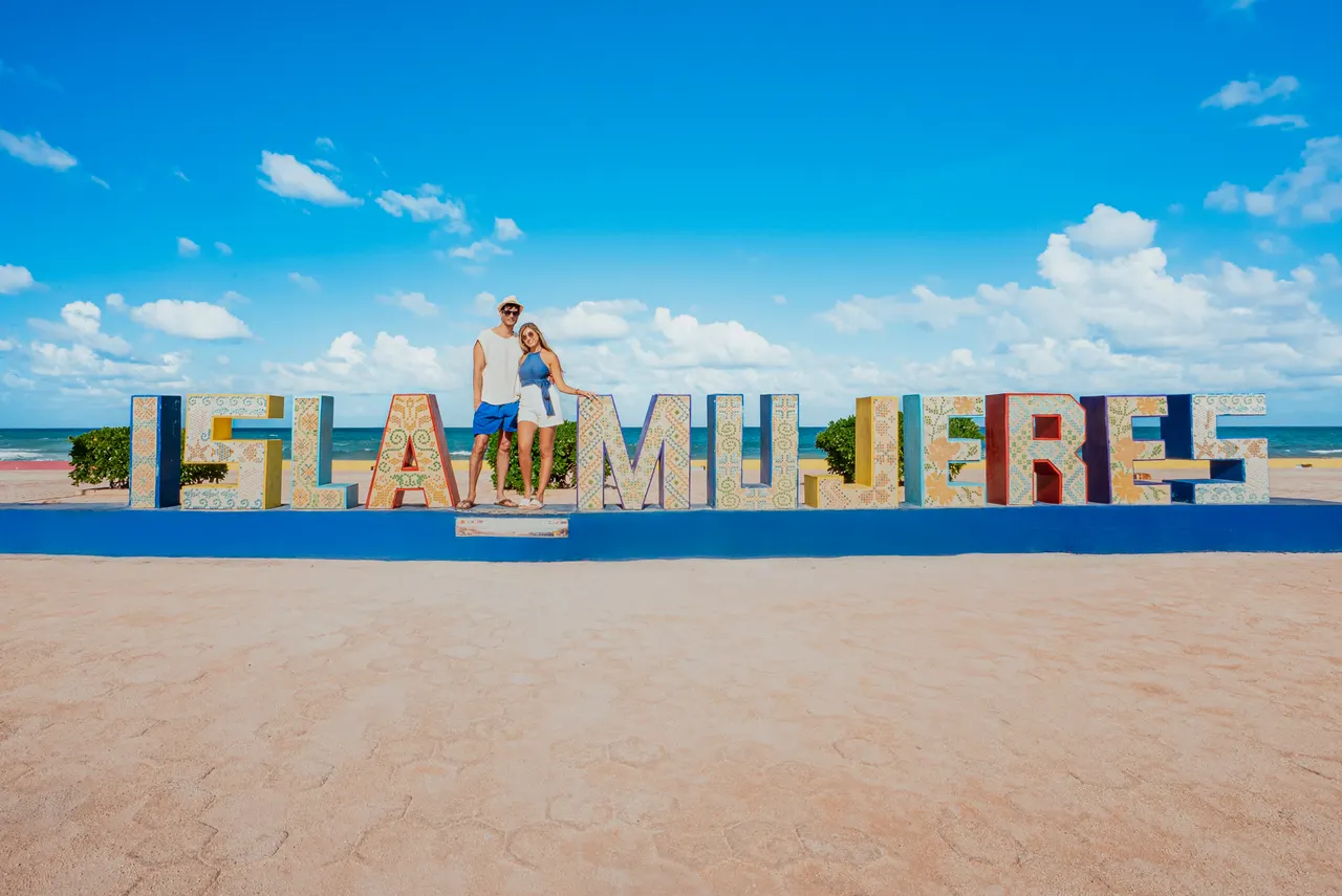 Pareja adulta joven posando junto a las letras gigantes 'ISLA MUJERES' en Playa Norte. El hombre viste shorts azules y la mujer sostiene un bolso blanco. De fondo, el mar turquesa del Caribe y un cielo azul despejado con nubes blancas. Punto icónico y fotogénico de la isla, perfecto para recuerdos de viaje.