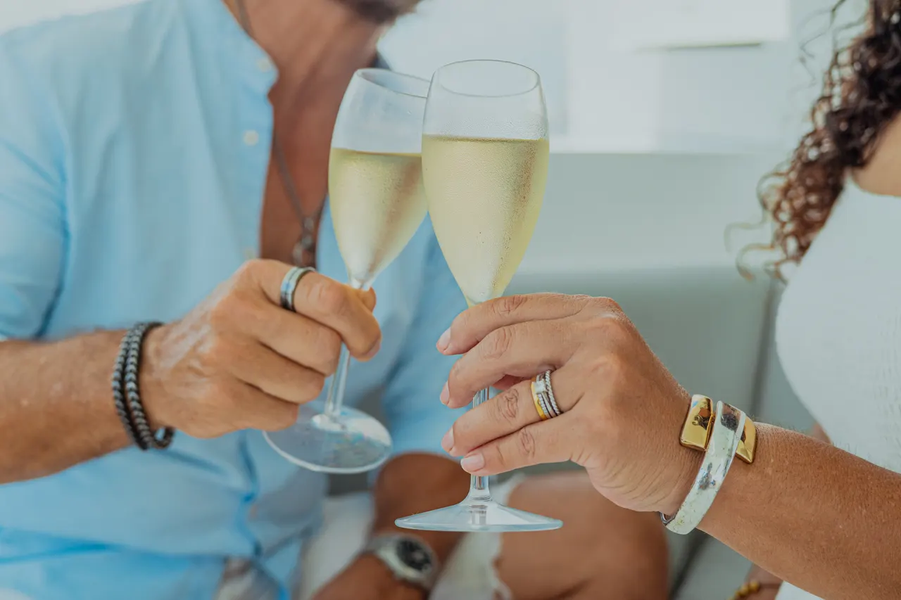 Pareja adulta brindando con copas de champagne espumoso sobre la cubierta de un catamarán. Ambos lucen anillos de boda y pulseras, con el mar turqueso visible de fondo. Ambiente romántico y celebratorio, perfecto para luna de miel o aniversario.