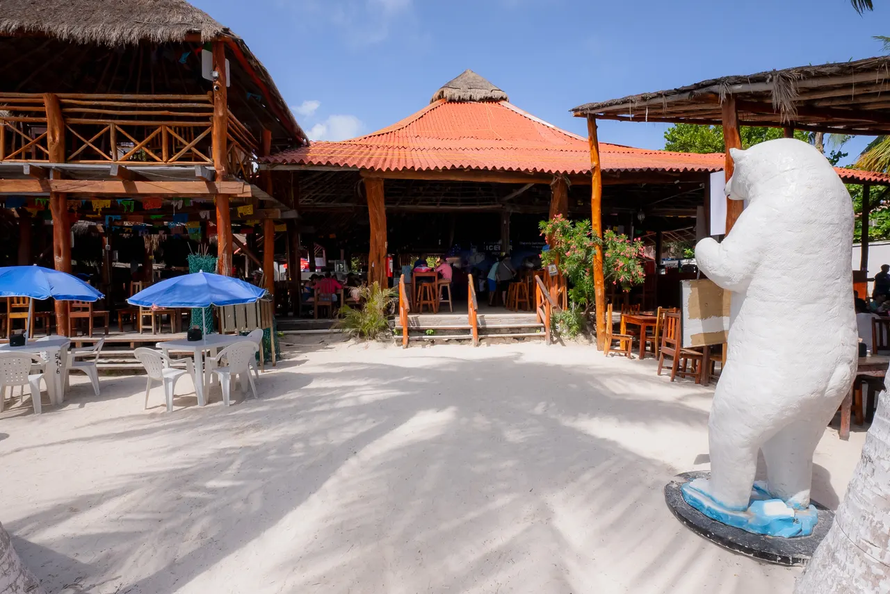 Instalaciones de un club de playa en Isla Mujeres con arquitectura rústica de madera y techos de palapa naranja. En primer plano, una estatua blanca decorativa; al fondo, palapas con sombrillas azules, mesas de madera y bar restaurante. Ambiente tropical, casual y acogedor bajo el cielo despejado.