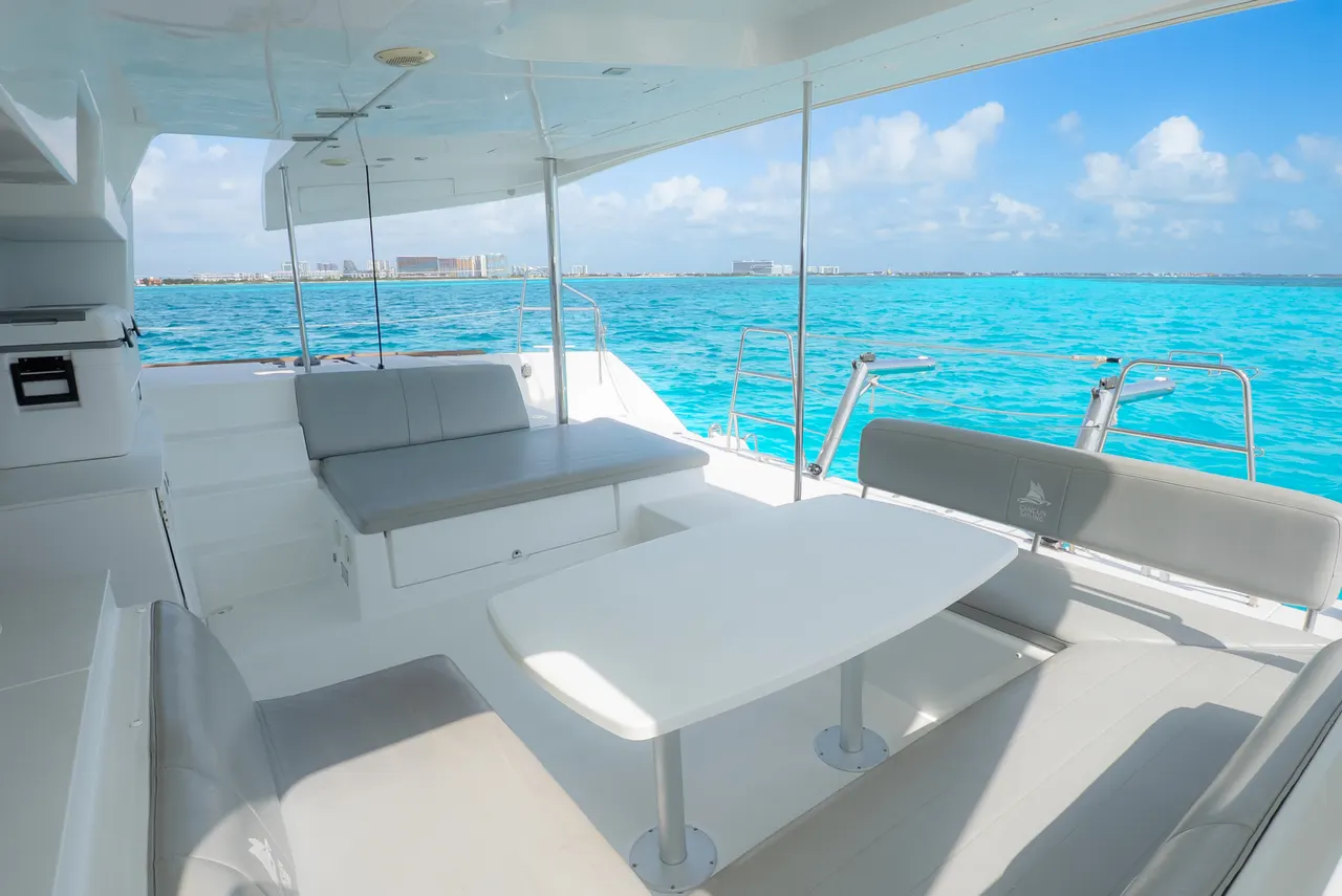 Vista de la cubierta principal de un catamarán moderno con asientos grises, mesas blancas y barandales de acero inoxidable. El agua turquesa del Caribe se extiende al horizonte con Cancún visible de fondo bajo un cielo azul despejado. Ambiente limpio, espacioso y premium.