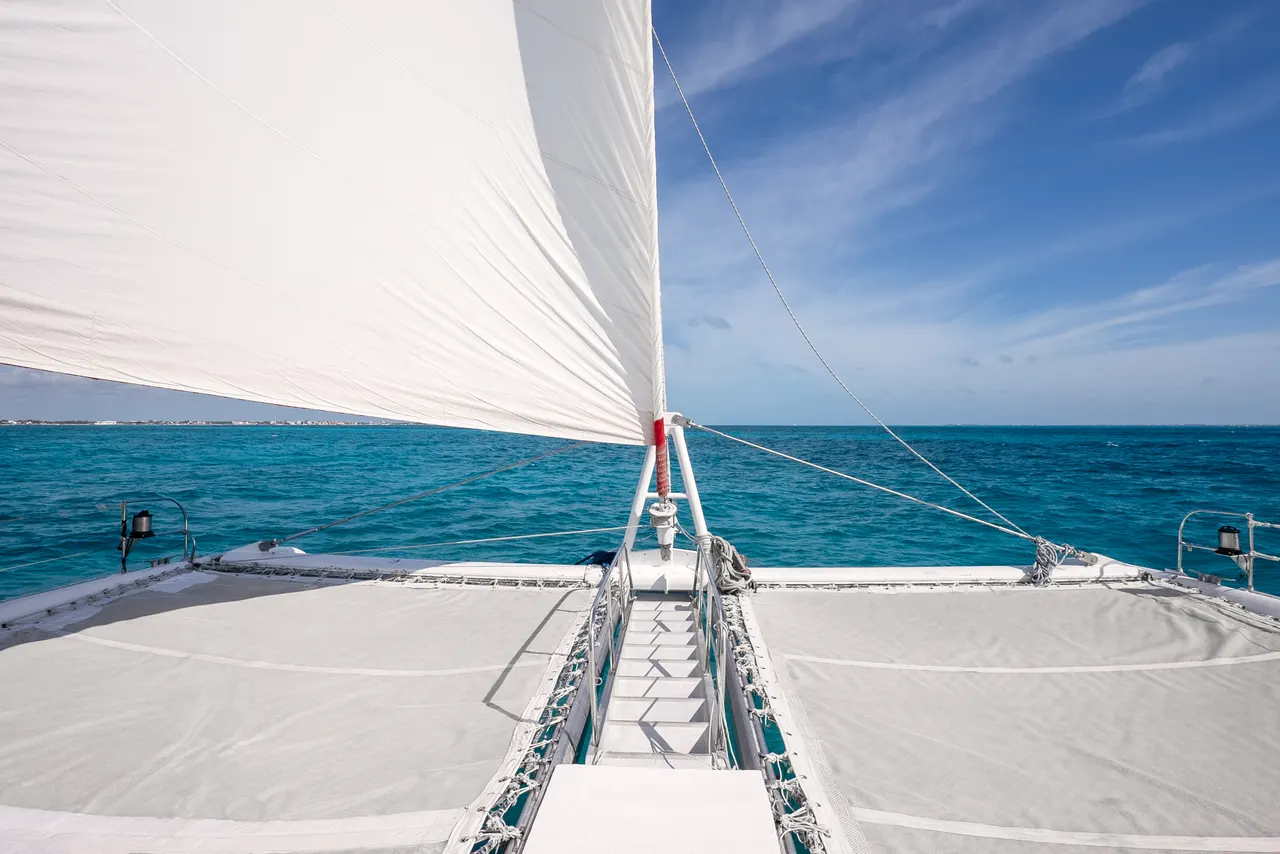Vista aérea cenital de la proa de un catamarán con vela blanca completamente desplegada, navegando sobre aguas turquesas cristalinas del Caribe. La composición muestra la red de proa, las velas tensas y el horizonte marino en condiciones de navegación perfectas.