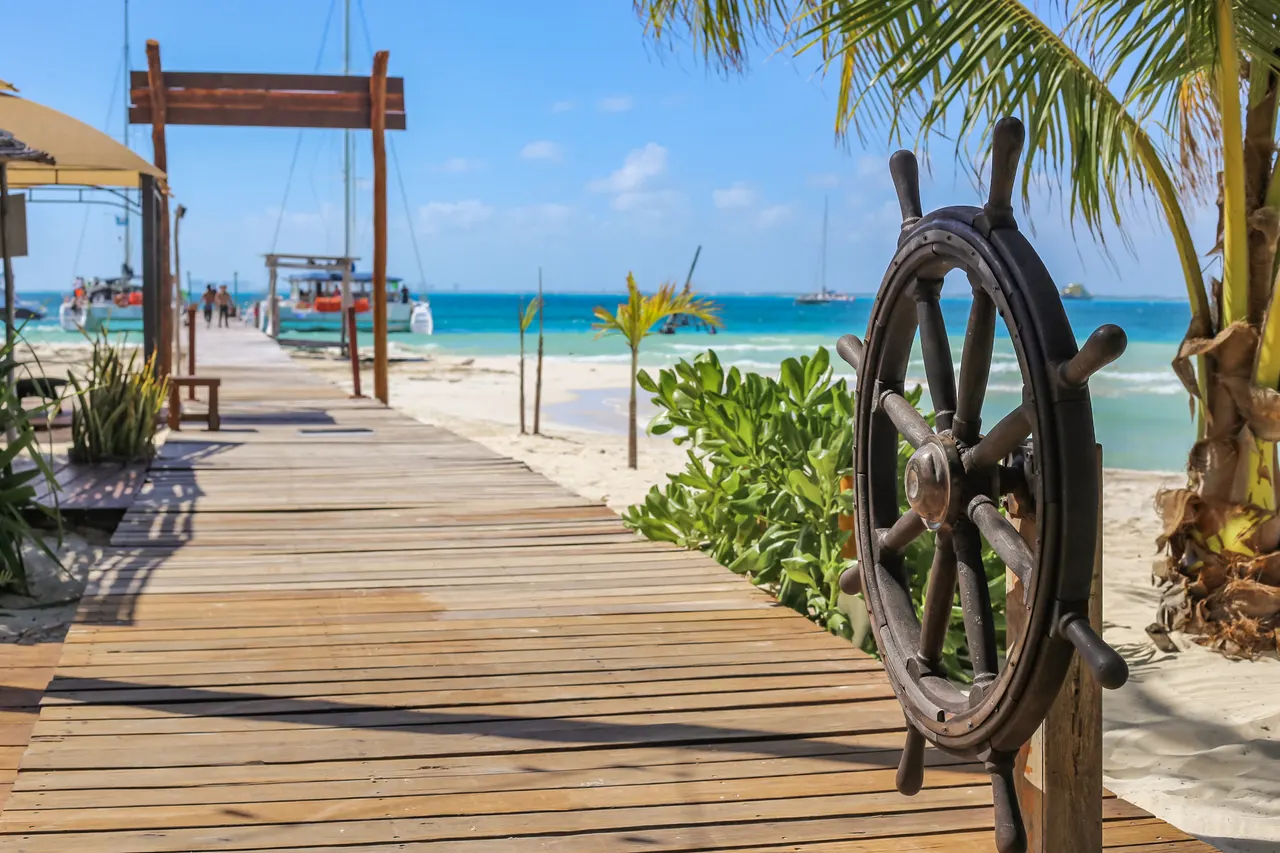 Primer plano de un timón de barco de madera oscura en un muelle de madera, con vistas al mar turquesa de Isla Mujeres. Al fondo, palmeras, playas de arena blanca, catamaranes anclados y bungalós, capturando la esencia tropical y náutica del destino.