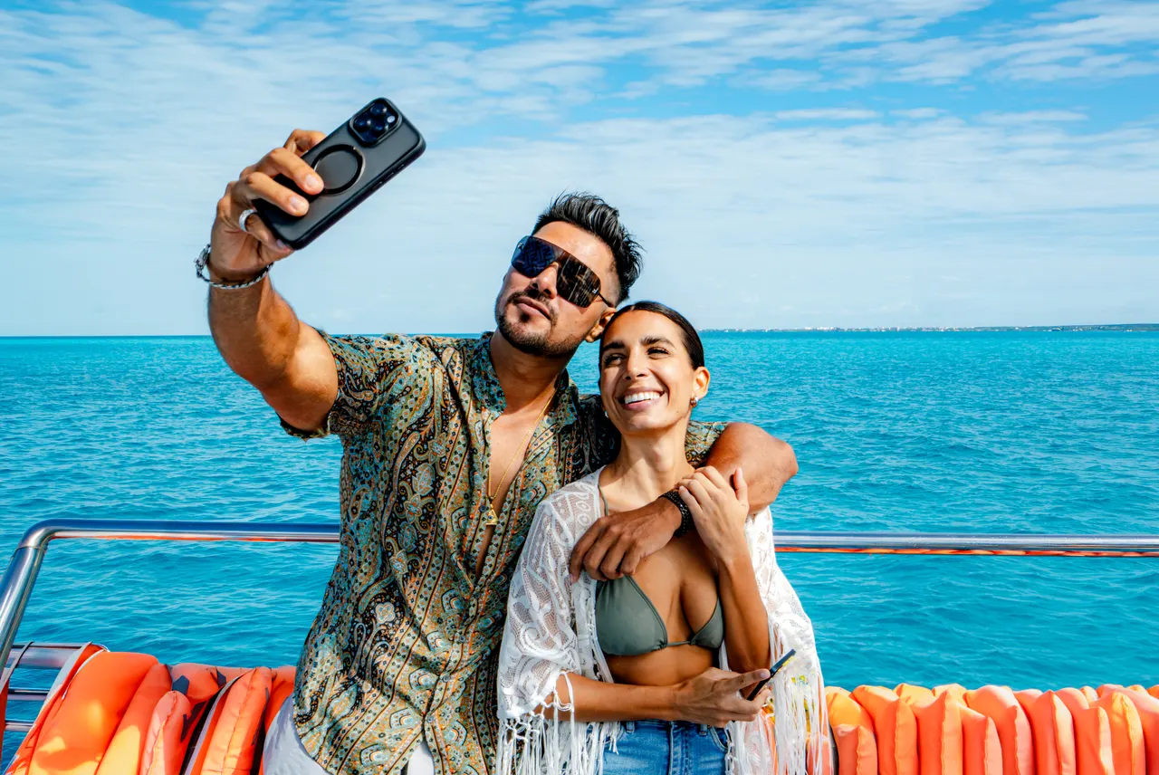 Pareja joven sonriente tomando un selfie a bordo de un catamarán, con el hombre levantando su teléfono hacia la cámara mientras abraza a la mujer. El fondo muestra el agua turquesa del Caribe bajo un cielo azul despejado, con las características boyas naranjas del barco visibles en primer plano. Ambiente casual, despreocupado y festivo.