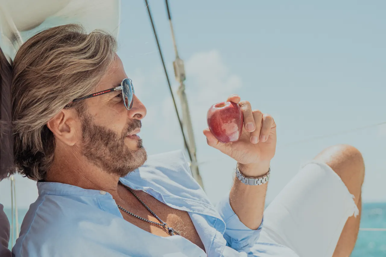 Hombre adulto con gafas de sol sosteniendo una manzana roja fresca en la cubierta de un catamarán, con el mar turquesa del Caribe de fondo. Ambiente casual y relajado, con vistas al horizonte marino claro.