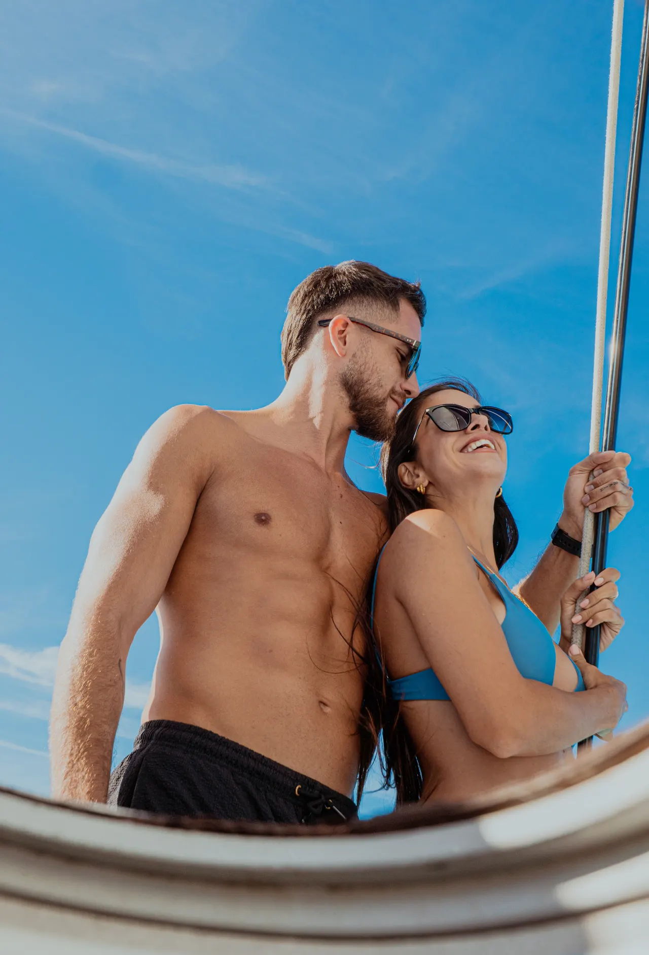 Pareja adulta en traje de baño disfrutando del sol en la cubierta de un catamarán, con el cielo azul caribeño de fondo. El hombre sostiene el mástil mientras abraza a la mujer, ambos sonríen y lucen relajados. Ambiente casual y romántico, captura la esencia de una escapada playera en el Caribe.