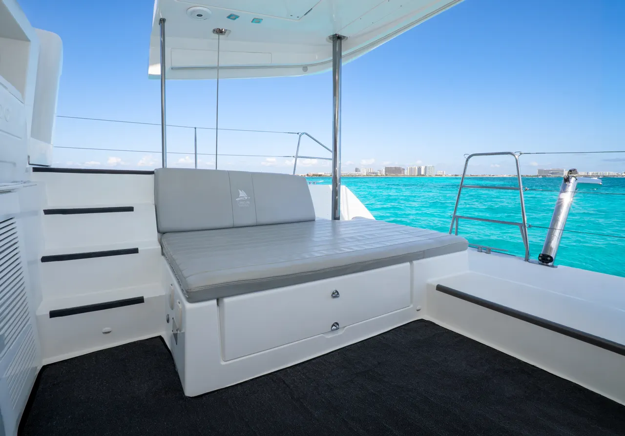 Interior de la cubierta de un catamarán de lujo con asientos grises y blancos, piso de madera clara y techo acristalado. Al fondo, aguas turquesas del Caribe y la costa de Cancún. Ambiente moderno, limpio y exclusivo con excelentes vistas panorámicas.