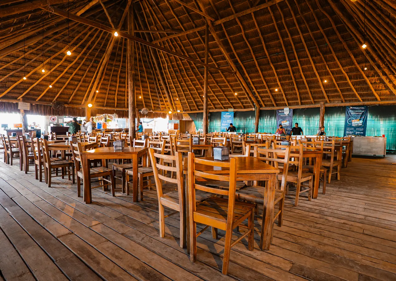 Interior de un restaurante de club de playa con estructura de palapa tradicional de madera, techo de paja y luces cálidas. Múltiples mesas de madera y sillas dispuestas para servir comidas, con vistas al mar turquesa a través de las aberturas. Personal de servicio visible al fondo, ambiente tranquilo y acogedor típico de instalaciones playeras en Cancún.