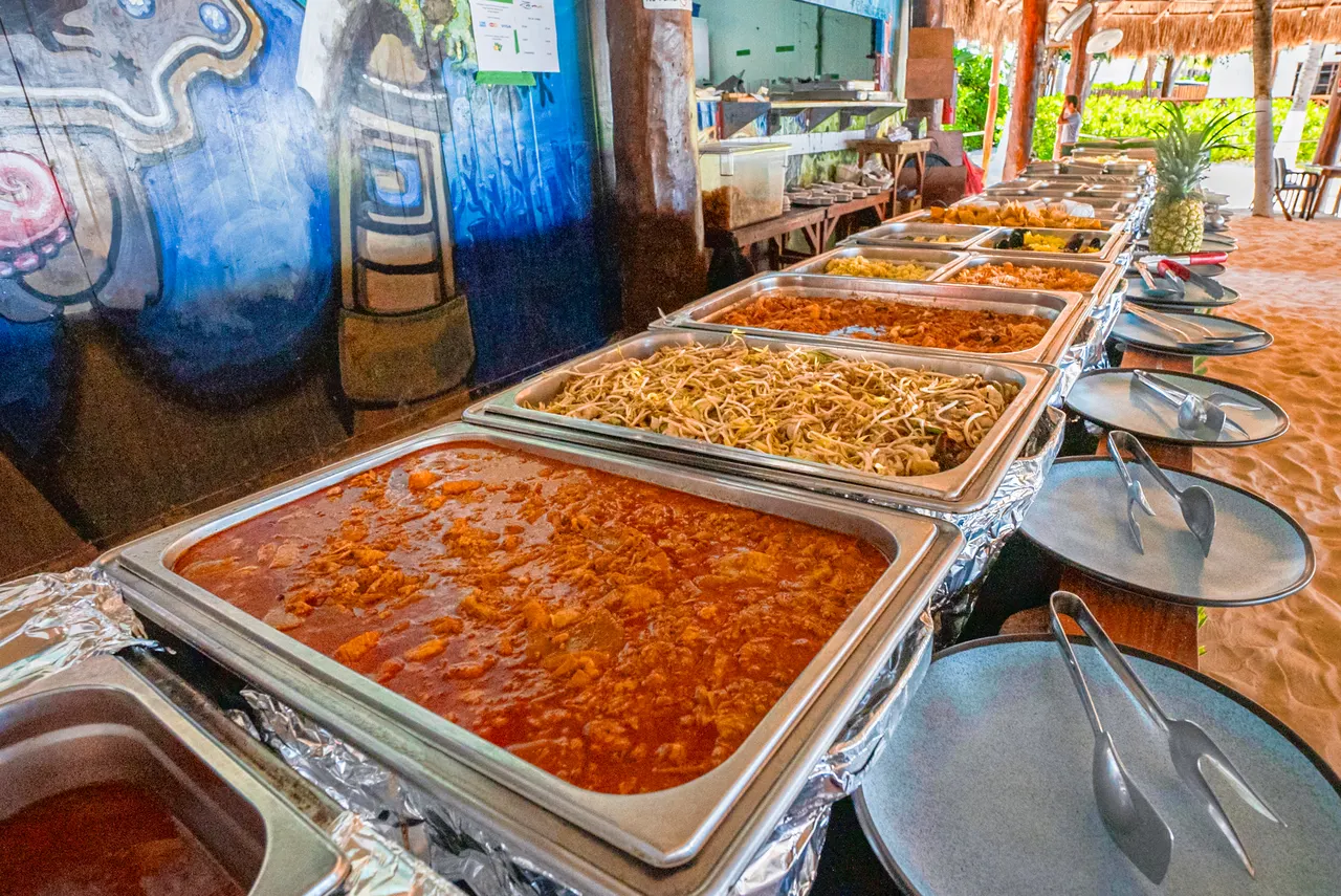 Buffet abundante con múltiples bandejas de acero inoxidable conteniendo platillos variados incluyendo arroces, guisos y proteínas. La escena se desarrolla en un club de playa con palapas de palma, decoración artística en la pared y ambiente tropical al aire libre, típico de las instalaciones de Isla Mujeres.
