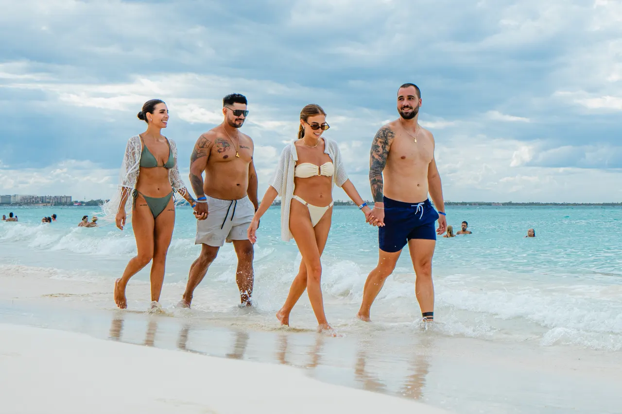 Grupo de cuatro adultos jóvenes caminando juntos en aguas poco profundas de una playa tropical con arena blanca y agua turquesa del Caribe. El ambiente es casual, divertido y relajado, con otros visitantes disfrutando al fondo. Ideal para promocionar experiencias de grupo y diversión en destinos de playa.