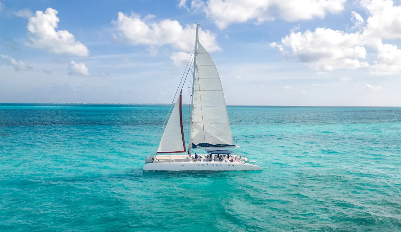 Catamarán de lujo navegando en aguas turquesas cristalinas del Caribe con velas blancas desplegadas. Pasajeros disfrutando a bordo bajo un cielo azul despejado con nubes blancas. La imagen captura la esencia de un tour marino en Cancún con el icónico color azul caribe de fondo.