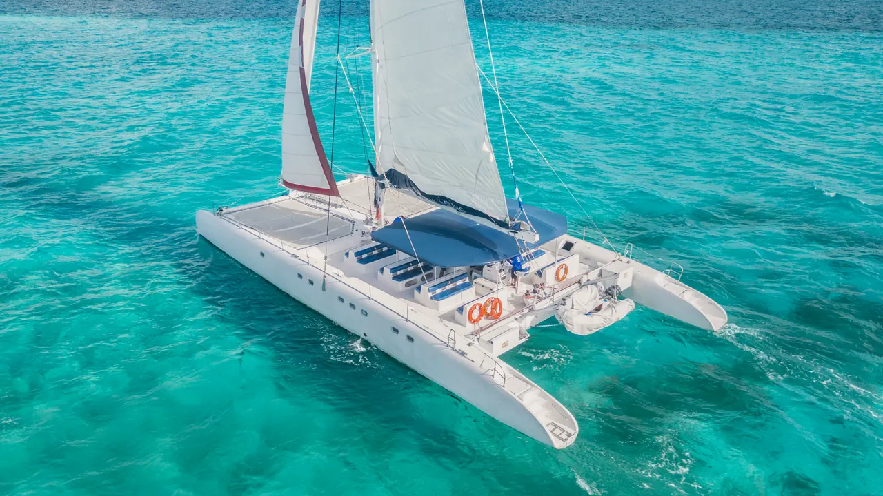 Vista aérea de un catamarán blanco con velas desplegadas navegando en las aguas cristalinas y turquesas del Caribe. El barco muestra su diseño elegante con cubierta espaciosa, velas blancas y rojas, y flotadores laterales, capturando la esencia del lujo y la aventura marina.