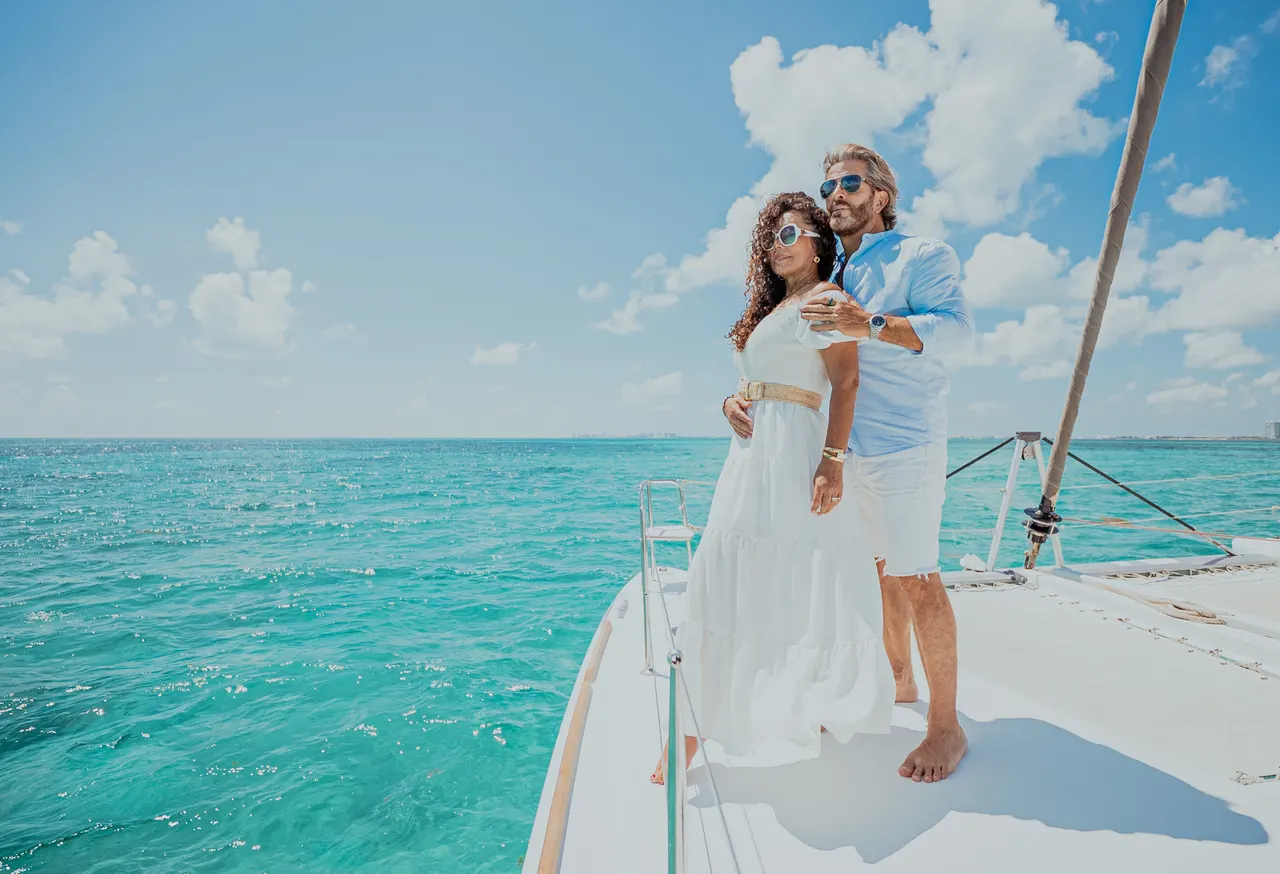 Pareja adulta posando en la cubierta frontal de un catamarán blanco, con el hombre abrazando a la mujer desde atrás. Ella viste un elegante vestido blanco fluido y él lleva camisa celeste, ambos con gafas de sol. El mar turquesa del Caribe brilla bajo un cielo azul con nubes blancas, creando una atmósfera romántica y exclusiva ideal para luna de miel o aniversario.