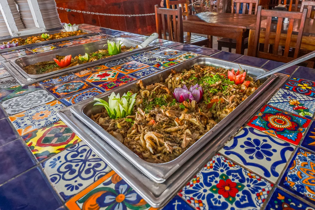 Buffet servido en charolas de acero inoxidable sobre mesas decoradas con azulejos talavera tradicionales mexicanos. Se aprecian carnes desmenuzadas, vegetales frescos como flores de cebolla y tomates, en un ambiente de restaurante rústico elegante con sillas de madera y detalles de lujo casual.