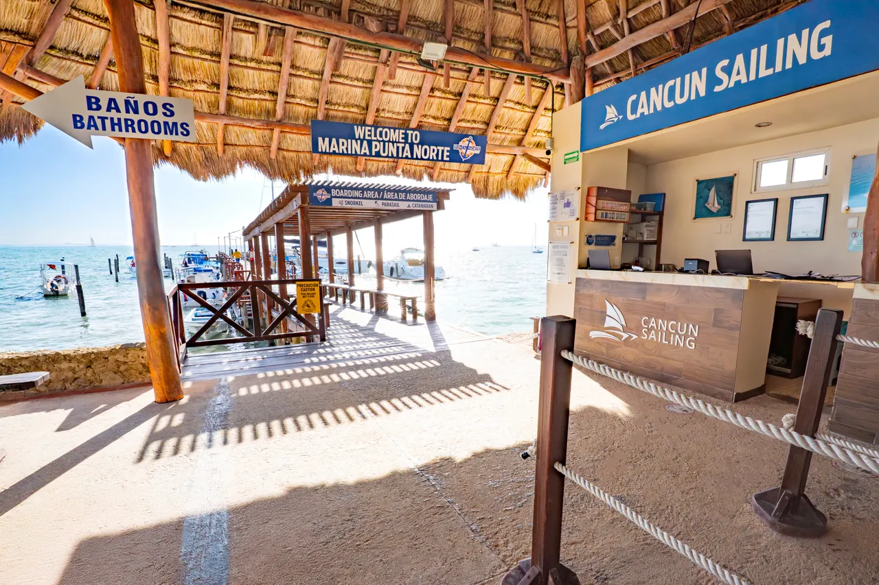 Instalaciones de Marina Punta Norte con palapas de techo de paja, mostrador de check-in de Cancun Sailing y vistas al mar turquesa. La marina cuenta con servicios de baños, zona de espera cubierta y acceso directo a los muelles donde están anclados los catamaranes. Ambiente profesional y acogedor para el embarque de pasajeros.