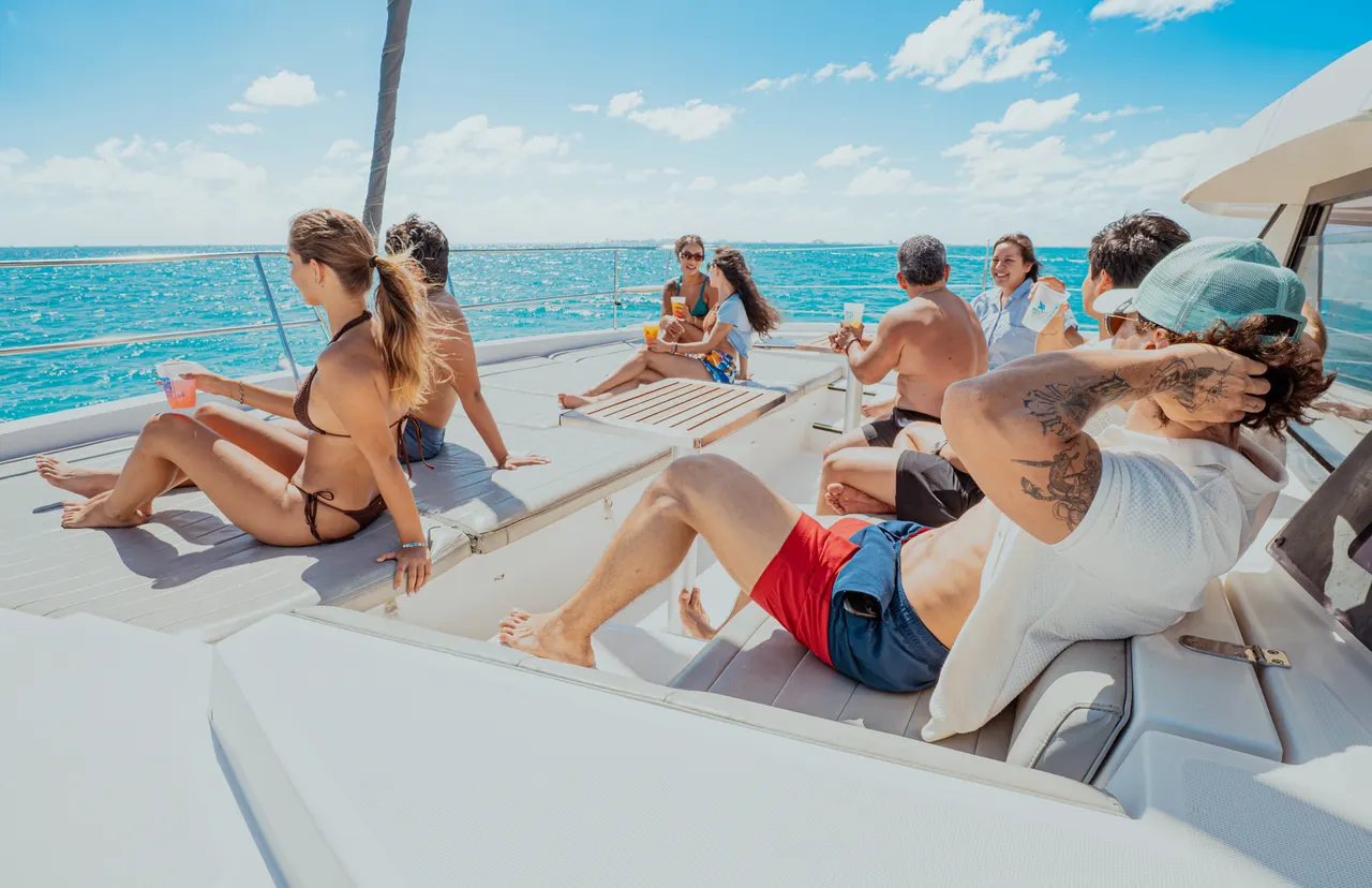 Grupo de adultos jóvenes relajándose en la cubierta de un catamarán bajo el sol, disfrutando del agua turquesa del Caribe. El ambiente es casual y festivo, con personas tomando bebidas, conversando y disfrutando del viaje. La composición muestra la amplitud de la cubierta y la belleza del entorno marino.
