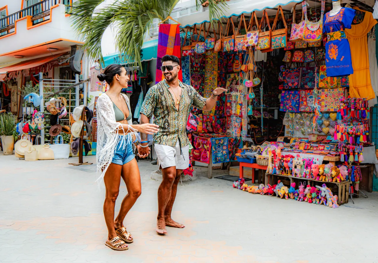 Pareja joven explorando las vibrantes tiendas de artesanía y souvenirs en Isla Mujeres, rodeados de coloridas bolsas tejidas, vestidos tradicionales y artículos turísticos. Ambiente casual y festivo con arquitectura típica caribeña y palmeras de fondo.