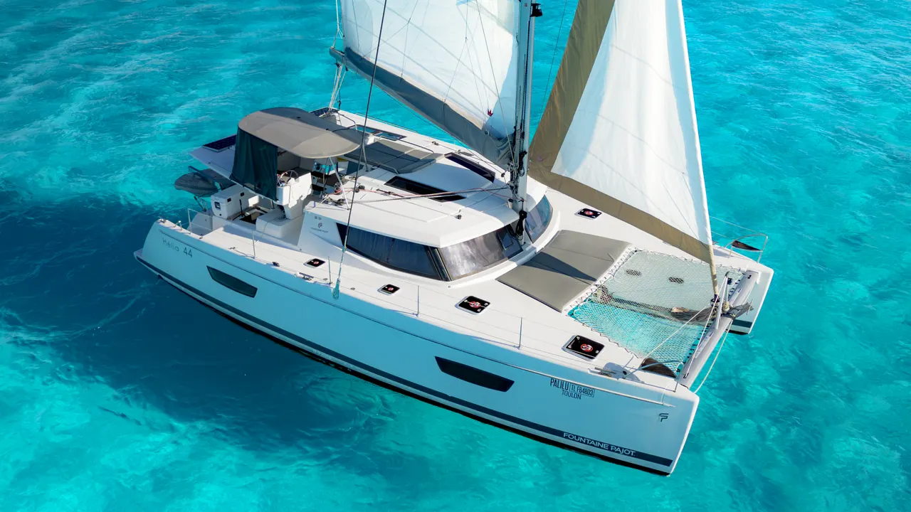 Vista aérea de un catamarán blanco de lujo con velas izadas navegando en las cristalinas aguas turquesas del Caribe. La embarcación muestra claramente su diseño moderno, cubierta espaciosa y red de proa, flotando en agua de color azul intenso. Imagen perfecta para promocionar la experiencia premium de navegación.