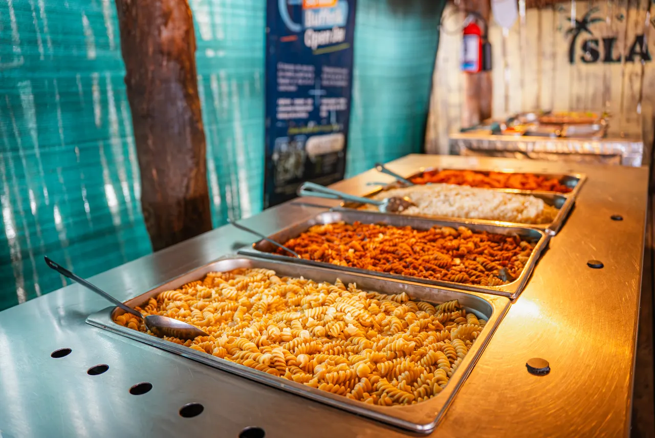 Mostrador de buffet con múltiples bandejas de acero inoxidable llenas de pastas de diferentes tipos y colores (fusilli rojo, blanco y integral) en un club de playa. Al fondo se ve la piscina turquesa y palapas, creando un ambiente de gastronomía casual y abundante ideal para disfrutar después del snorkel.