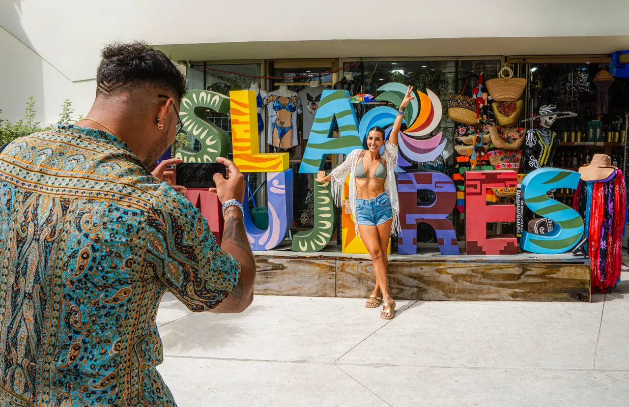 Turista con camisa estampada toma una foto con su smartphone frente a la icónica letra 'ISLA MUJERES' en colores vibrantes. Una mujer joven posa en bikini frente a la tienda de souvenirs, rodeada de accesorios playeros, sombreros y artesanías típicas de la isla. Ambiente turístico casual y vibrante en una calle de Isla Mujeres.